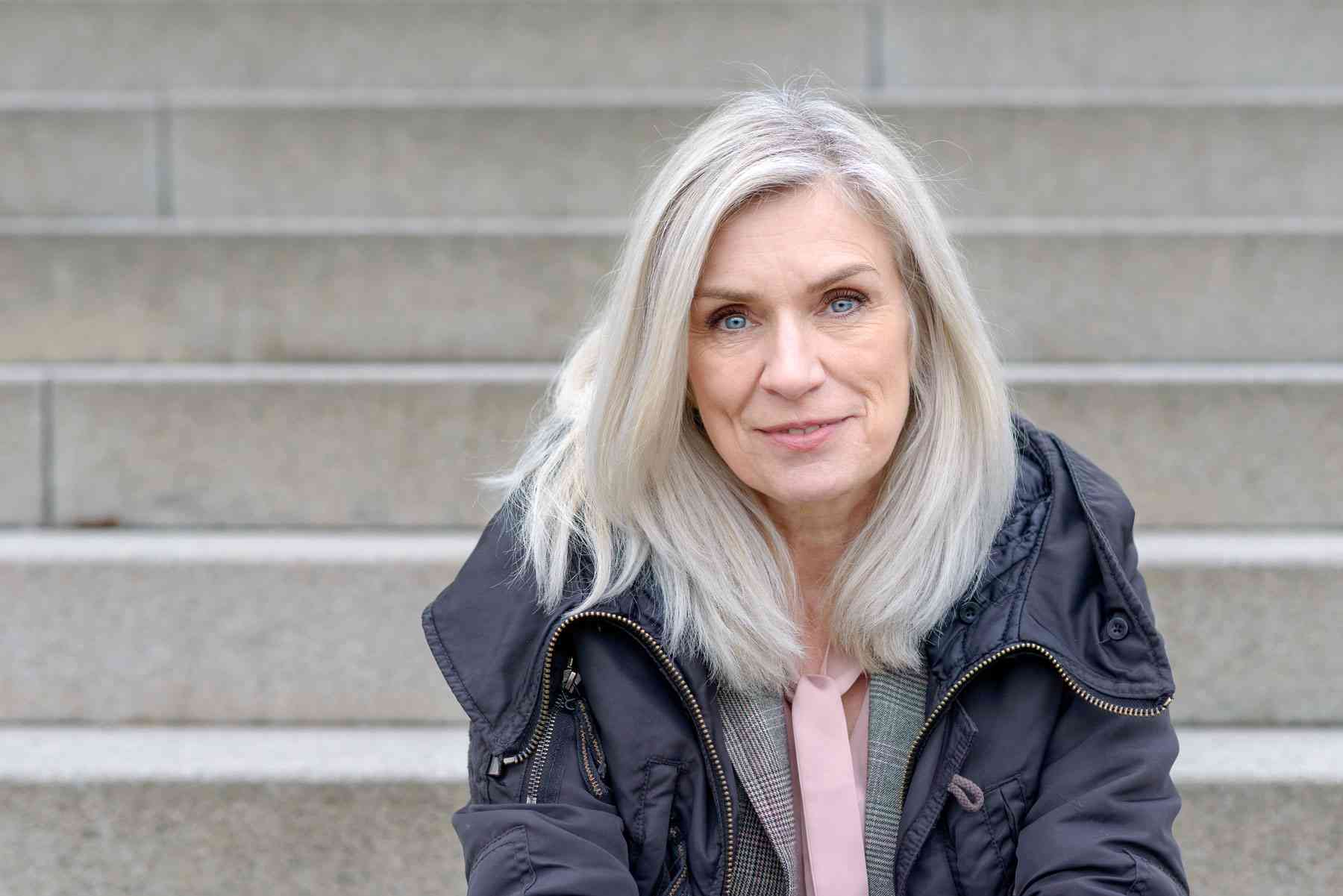 Ældre kvinde med langt gråt hår og blå øjne sidder på en stentrappe iført en mørk jakke og skjorte med slipsedetalje.