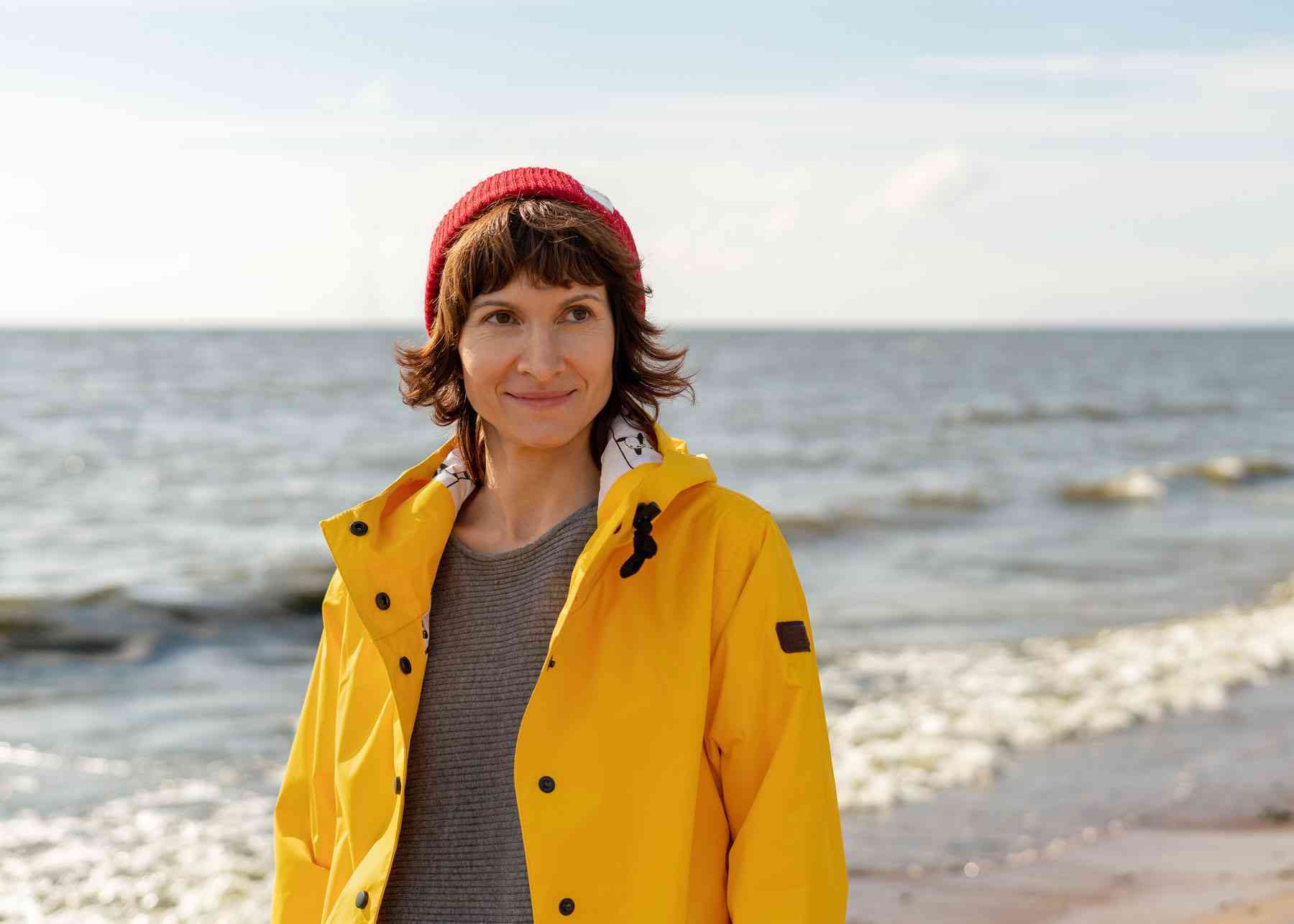 Kvinde med kort brunt hår iført en gul regnjakke og rød hue står på en strand foran havet en solrig dag.