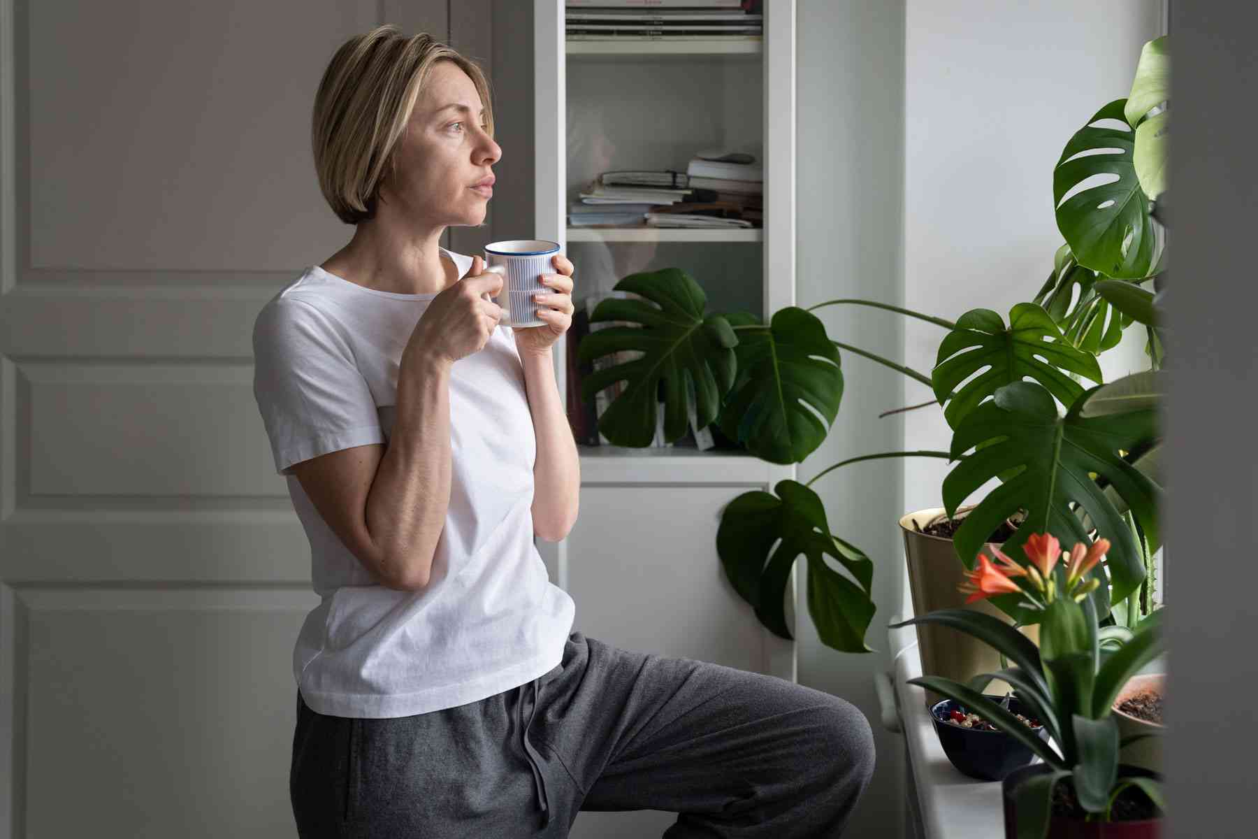 Kvinde i hvid T-shirt og grå bukser står ved et vindue med grønne planter og holder en kop, mens hun ser ud i dagslyset.